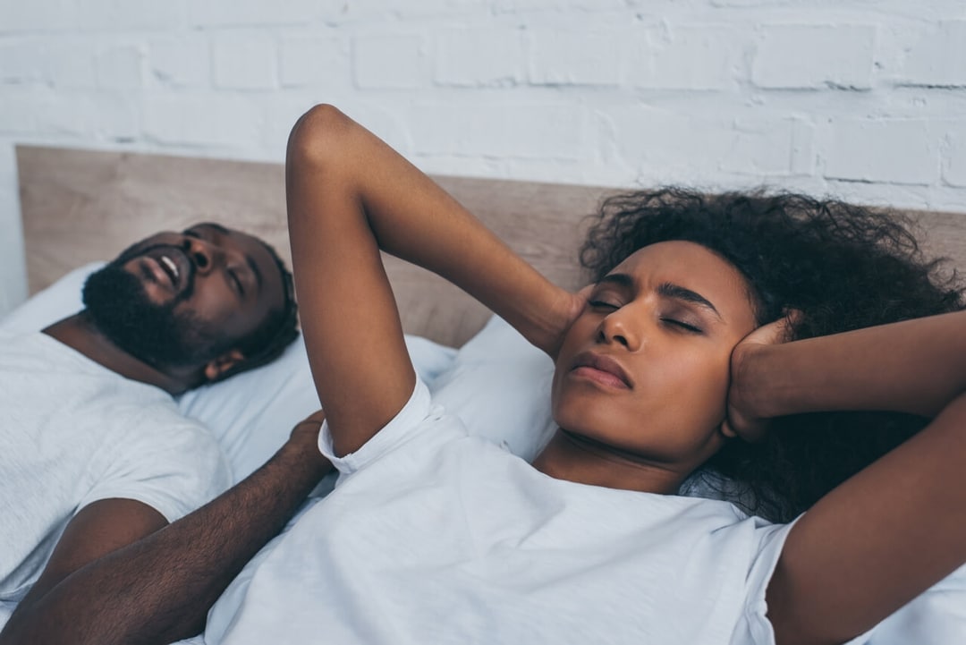 A man snoring in bed next to his partner who's holding her ears.
