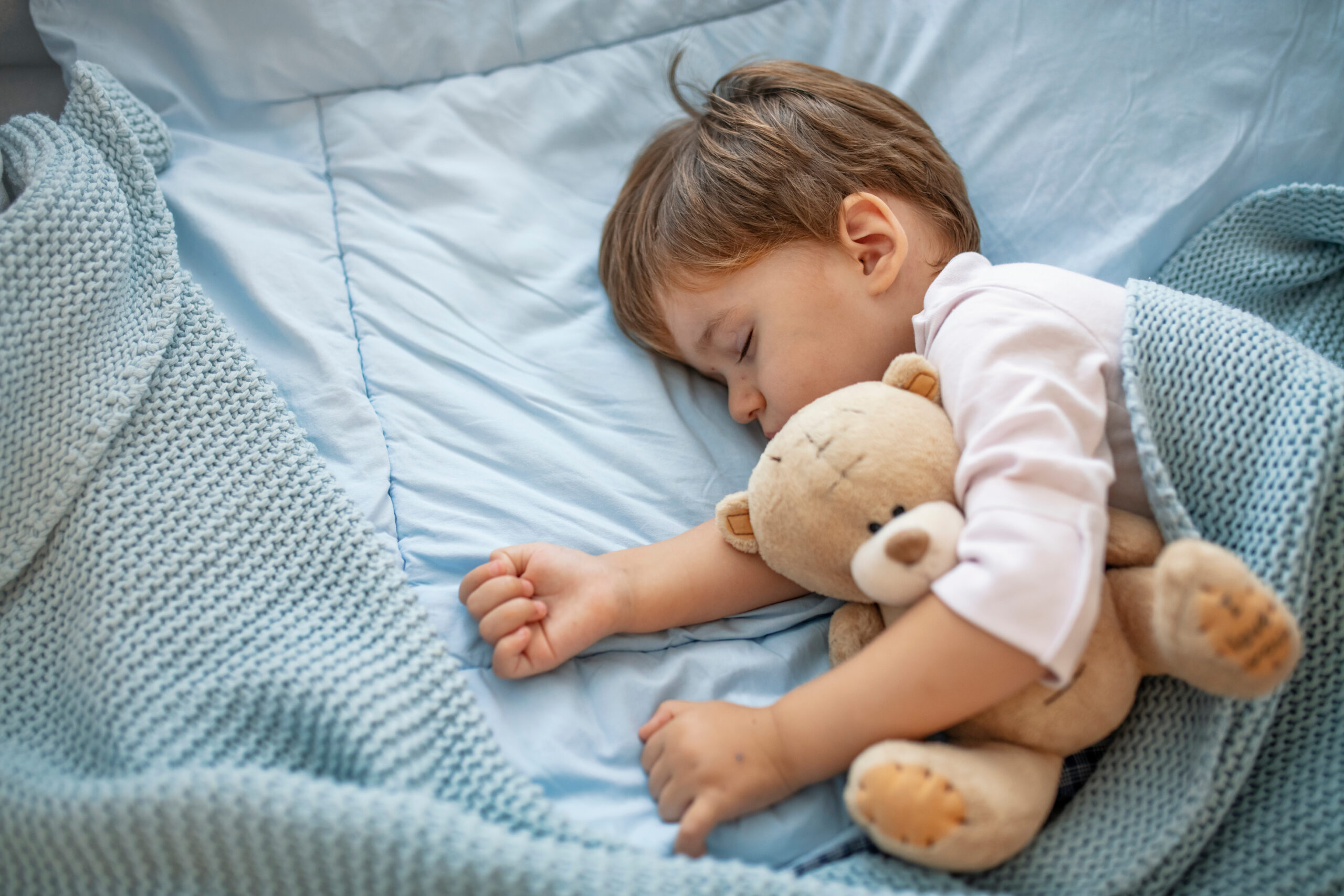 Healthy child, sweetest blonde toddler boy sleeping in bed holding her teddy bear. Adorable toddler girl taking a nap in a grey bed holding her teddy bear. Baby toddler asleep with teddy bear
