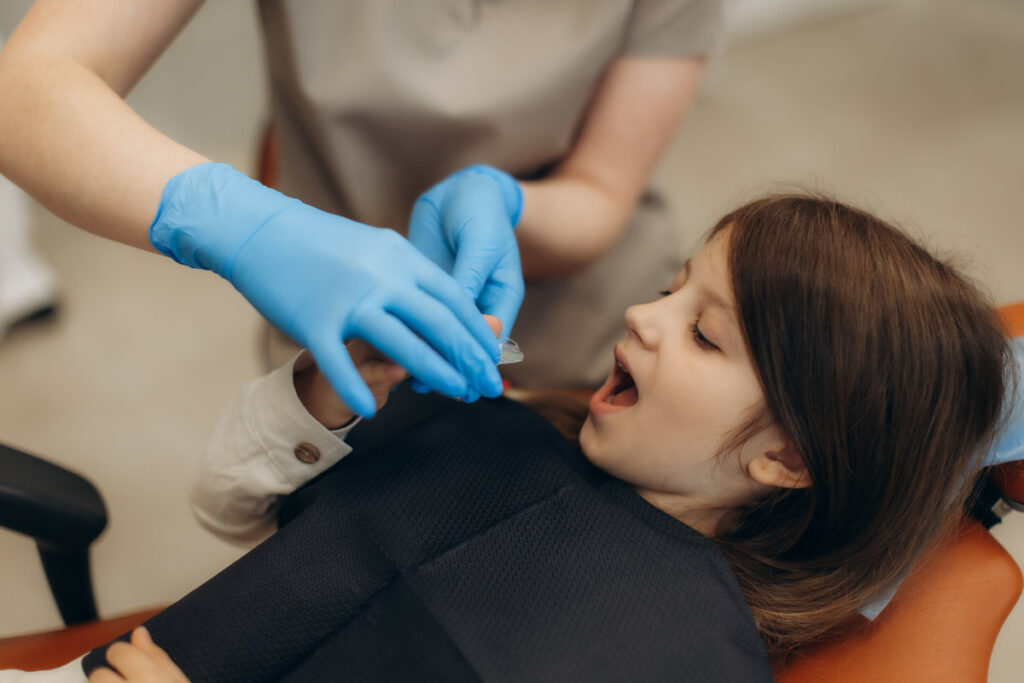 Pediatric dentist applying fluoride varnish on teeth of little girl in dental clinic, preventive dentistry and dental care concept